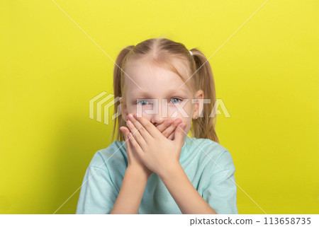 A beautiful seven-year-old girl covers her mouth with her hands on a yellow background. Concept of fear of dentists and dental treatment. Dentophobia in children. A beautiful seven-year-old girl covers her mouth with her hands on a yellow background. Concept of fear of dentists and dental treatment. Dentophobia in children. 113658735