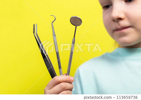 A beautiful little girl aged seven years old holds dental instruments on a yellow background. The concept of treatment of periodontal diseases, intraosseous implantation with subsequent prosthetics. A beautiful little girl aged seven years old holds dental instruments on a yellow background. The concept of treatment of periodontal diseases, intraosseous implantation with subsequent prosthetics. 113658736