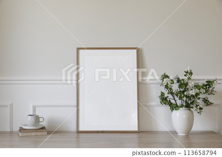 Springtime breakfast still life. Empty picture frame poster mockup. White ceramic vase with blooming apple tree branches. Cup of coffee, tea on wooden table, desk with old books. Scandi home interior 113658794