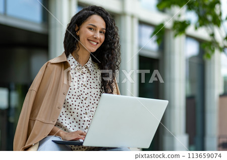 A smiling Hispanic woman uses her laptop while seated outdoors. Dressed in a stylish trench coat, she works amidst modern urban settings. 113659074