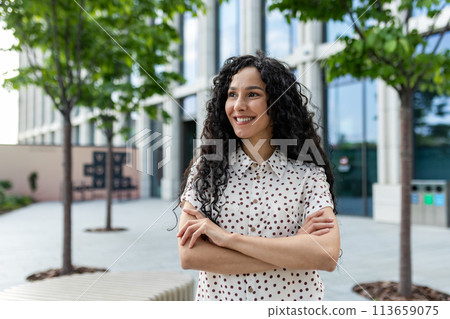 Young happy hispanic female student with arms crossed outside university campus smiling contentedly and looking away. 113659075