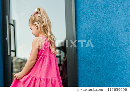 Child girl with hearing aid and cochlear implantation system. Kid with hearing problems walks outdoor in summer. Inclusion and diversity concept. Back view copy space 113659606