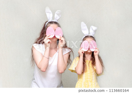 Two girls with Easter bunny ears close their eyes with Easter eggs. Easter is fun. Copy space. Two girls with Easter bunny ears close their eyes with Easter eggs. Easter is fun. Copy space. 113660164