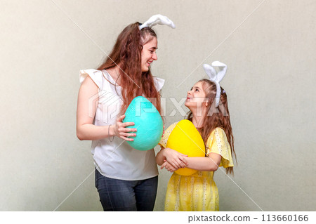 Two girls wearing Easter bunny ears look at each other and hold Easter eggs in their hands. Two girls wearing Easter bunny ears look at each other and hold Easter eggs in their hands. 113660166