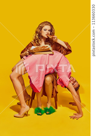Flexible woman stands in bridge gymnastic pose as table for pretty woman who eating pizza on her against yellow studio background. Flexible woman stands in bridge gymnastic pose as table for pretty woman who eating pizza on her against yellow studio background. 113660330