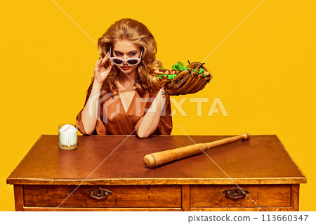 Stylish woman with sunglasses holds hotdog in baseball glove, with bat at wooden desk, against yellow studio background. 113660347