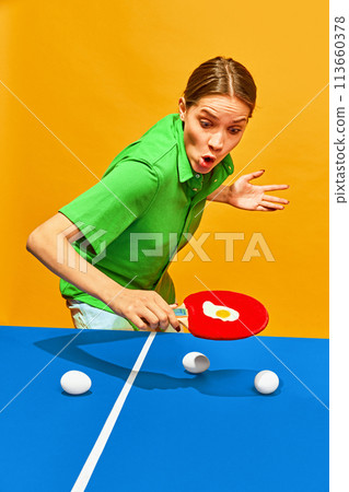 Woman with surprise facial expression playing tennis with fried egg on racquet against yellow studio background. Woman with surprise facial expression playing tennis with fried egg on racquet against yellow studio background. 113660378