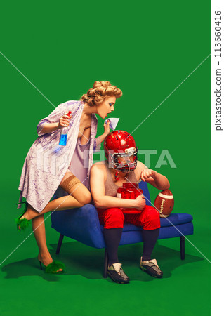 Elegant woman in silky bathrobe cleaning helmet on her husband, athlete, who watching an American football match on television 113660416