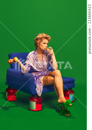 Girl drinks cocktail while sitting on vintage chair that has jars of vitamins instead of legs against vibrant green background. Girl drinks cocktail while sitting on vintage chair that has jars of vitamins instead of legs against vibrant green background. 113660423