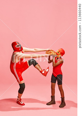 One wrestler in high kick pose holding pizza, other in mask eagerly receiving it against pink studio background. One wrestler in high kick pose holding pizza, other in mask eagerly receiving it against pink studio background. 113660440