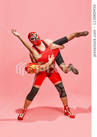 Athlete in red costume with mask executing wrestling move against partner dressed same costume against pink studio background. Athlete in red costume with mask executing wrestling move against partner dressed same costume against pink studio background. 113660456