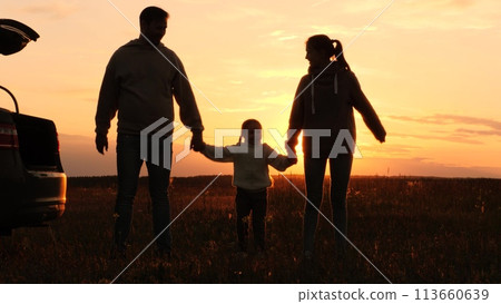Family holding hands at sunset, silhouettes near car. Joint recreation in nature. Mother father daughter in orange setting sun in field. Family picnic road trip, parents kid enjoy each other's company Family holding hands at sunset, silhouettes near car. Joint recreation in nature. Mother father daughter in orange setting sun in field. Family picnic road trip, parents kid enjoy each other's company 113660639