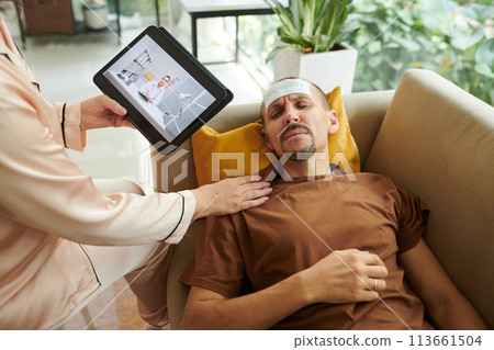 Wife video calling doctor when husband lying on couch with cooling patch on forehead 113661504