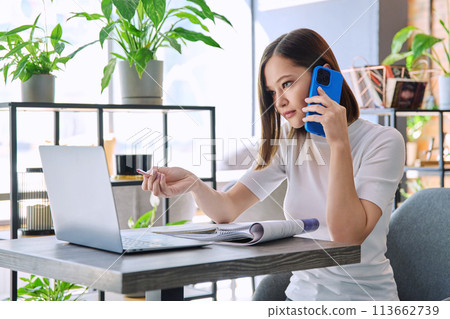 Young woman sitting at table with laptop talking on mobile phone in coworking Young woman sitting at table with laptop talking on mobile phone in coworking 113662739