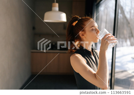 Side view of athletic young woman in sport outfit standing by window and drinking fresh water in morning after fitness training workout exercise at home, enjoying sunny weather and lifestyle. Side view of athletic young woman in sport outfit standing by window and drinking fresh water in morning after fitness training workout exercise at home, enjoying sunny weather and lifestyle. 113663591