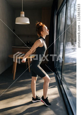 Side view of sporty slim young woman in outfit preforming warming up exercise, stretching ankle and arm muscles before workout, standing by window with beautiful snowy winter nature view on sunny day 113663633