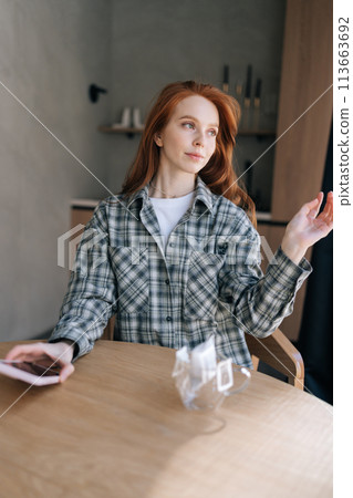 Vertical portrait of charming young woman sitting at table with drip coffee, holding in hands mobile phone in kitchen at home. Beautiful redhead female in casual wear drinking hot coffee alone. 113663692