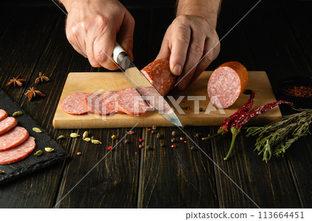 Slicing meat sausage. Concept of preparing or serving a sausage dish for a snack in a street restaurant. Knife in the hand of a cook Slicing meat sausage. Concept of preparing or serving a sausage dish for a snack in a street restaurant. Knife in the hand of a cook 113664451