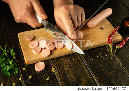 Cooking a delicious breakfast with milk sausages by the hands of a chef on the kitchen table. Cutting vienna sausage into small pieces with a knife Cooking a delicious breakfast with milk sausages by the hands of a chef on the kitchen table. Cutting vienna sausage into small pieces with a knife 113664457