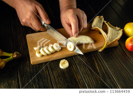 Finely sliced banana on a kitchen board. Cooking a fruit dish on the kitchen table with the hands of a chef Finely sliced banana on a kitchen board. Cooking a fruit dish on the kitchen table with the hands of a chef 113664459