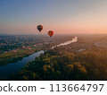 Two colorful air balloons flying over green park and river in small european city at summer sunrise 113664797