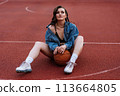 Outdoor portrait of dreaming girl in bra and jacket posing on the ground. SWAG concept. Excited brunette woman sitting at basketball court with ball between her legs. 113664805