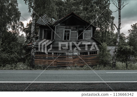 An old wooden abandoned and dilapidated leaning house after a fire at dusk next to the road in the foreground. Haunted house. An old wooden abandoned and dilapidated leaning house after a fire at dusk next to the road in the foreground. Haunted house. 113666342