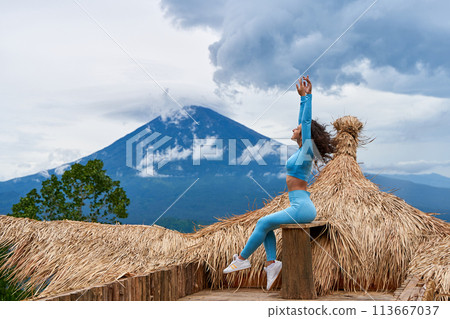 A young woman poses on a large straw photo zone as a bird on a viewing platform and enjoys the view of the sacred Mount Agung volcano hidden by clouds on a rainy day on the island of Bali. A young woman poses on a large straw photo zone as a bird on a viewing platform and enjoys the view of the sacred Mount Agung volcano hidden by clouds on a rainy day on the island of Bali. 113667037