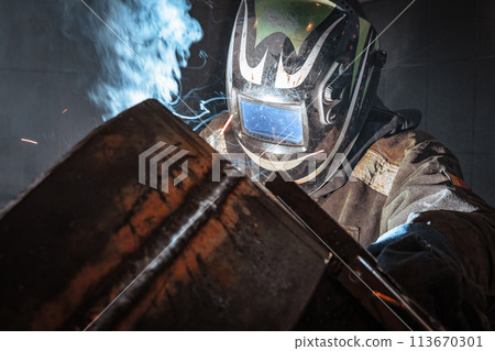 A man wearing a welding helmet is working on a piece of metal. Concept of danger and risk, as the A man wearing a welding helmet is working on a piece of metal. Concept of danger and risk, as the 113670301