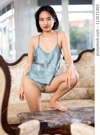 In cozy living room, naked Asian woman in nightie stands by sofa 113671068
