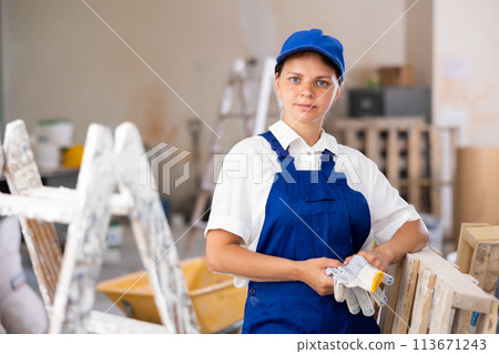 Portrait of positive builder woman in blue overalls next to stepladder 113671243