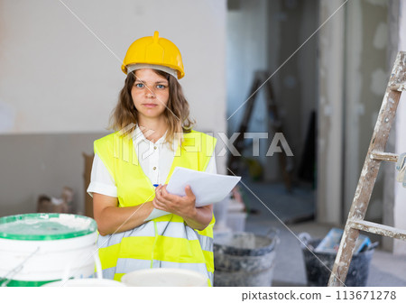 Woman foreman in a protective helmet and yellow vest checks the completed construction work on drawing Woman foreman in a protective helmet and yellow vest checks the completed construction work on drawing 113671278