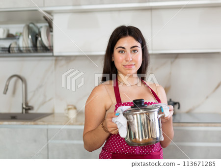 Young woman in apron posing with saucepan in kitchen 113671285