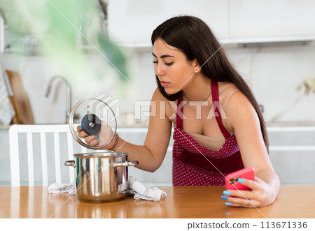 Young woman in apron with phone looks at pan 113671336