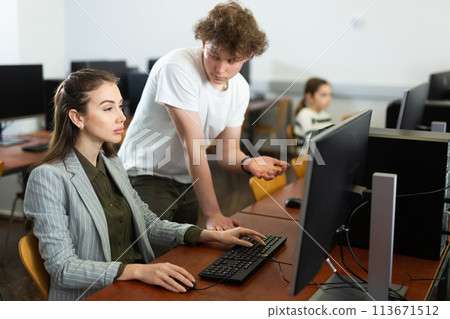 Teenage student talking to female teacher sitting at computer in class Teenage student talking to female teacher sitting at computer in class 113671512