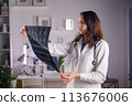 A young adult white female doctor in a medical mask and a white coat holds a CT scan in her hands and looks at it carefully 113676006