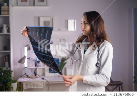 A young adult white female doctor in a medical mask and a white coat holds a CT scan in her hands and looks at it carefully A young adult white female doctor in a medical mask and a white coat holds a CT scan in her hands and looks at it carefully 113676006