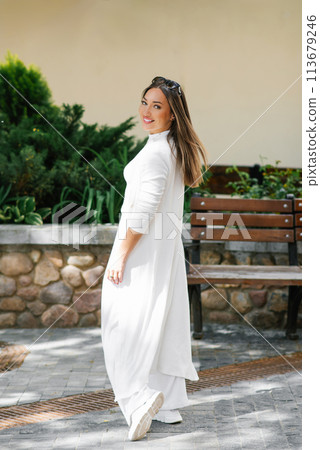 Happy woman in a white dress stands in front of a bench at city 113679246
