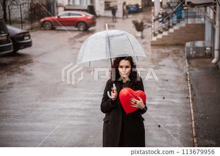 A woman holding a red heart under a clear umbrella. The umbrella is open and the woman is standing in the rain. A woman holding a red heart under a clear umbrella. The umbrella is open and the woman is standing in the rain. 113679956