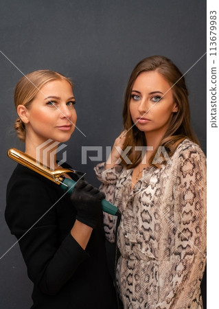 Two women are posing for a photo, one holding a hair curler. Scene is casual and friendly, as the women are smiling and looking at the camera. 113679983