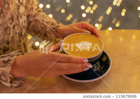 Close up cup of latte coffee in a coffee shop. Female hands hold a cup of coffee with foam latte in the shape of a heart on a table in a cafe. On the background, the lights are in blur. 113679986