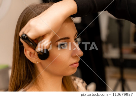 Makeup beauty salon. Makeup artist applies powder and blush. Beautiful female face. Makeup master's hand puts blush on cheeks of beauty model girl. Makeup in progress 113679987