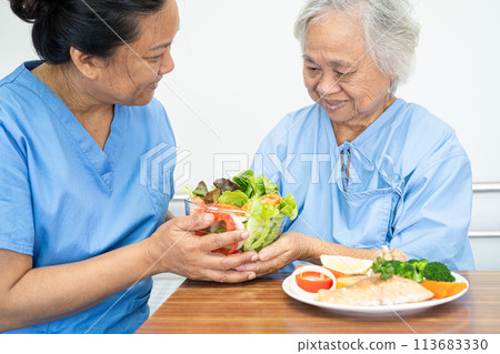Asian elderly woman patient eating Salmon steak breakfast with vegetable healthy food while sitting and hungry on bed in hospital. 113683330