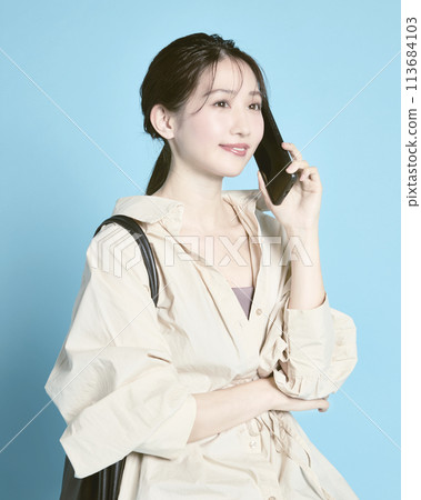 一位年輕女子拿著智慧型手機和一個包包，在藍色背景下拍照 113684103