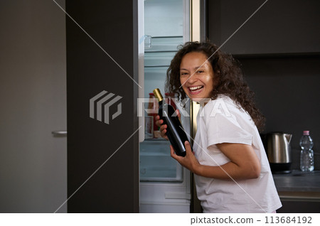 Alcoholism, mental health and social problems. Young curly haired brunette woman with alcohol addiction, holding a bottle of wine and drinking alcohol in the morning, smiling looking at camera. 113684192