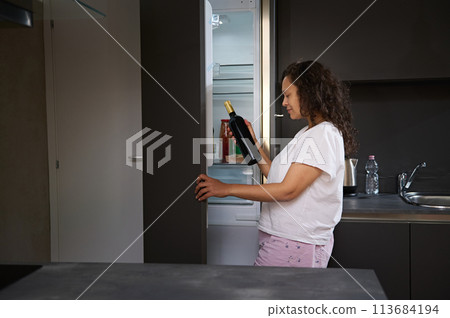Woman suffers from dipsomania, taking out a bottle of wine from the fridge at home kitchen. Female with alcohol addiction drinking alcohol in the morning. Alcoholism, mental health and social problems 113684194