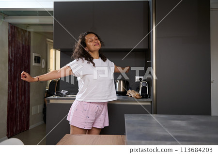 Portrait of a multi ethnic happy curly haired young woman in pajamas, stretching and smiling standing in minimalist modern kitchen. Cheerful female stretching and waking up early in morning at home Portrait of a multi ethnic happy curly haired young woman in pajamas, stretching and smiling standing in minimalist modern kitchen. Cheerful female stretching and waking up early in morning at home 113684203