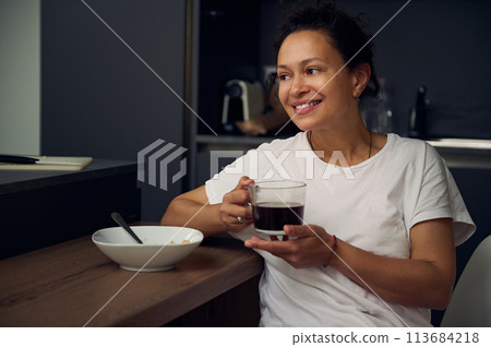 Portrait of joyful young woman enjoying a cup of coffee at home. Attractive pretty lady drinking hot freshly brewed coffee, smiling looking away, sitting at kitchen table while taking a breakfast Portrait of joyful young woman enjoying a cup of coffee at home. Attractive pretty lady drinking hot freshly brewed coffee, smiling looking away, sitting at kitchen table while taking a breakfast 113684218