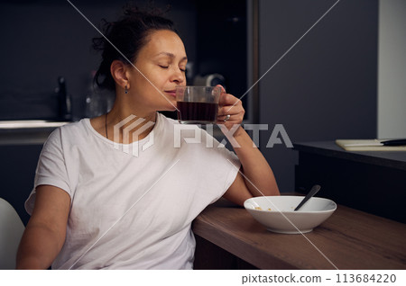 Young pretty woman in white pajamas, sitting with her eyes closed at the kitchen table, taking sip of a freshly brewed espresso coffee. Start your day with energizing freshly brewed coffee 113684220