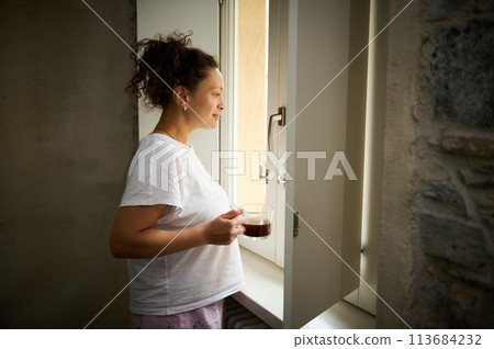 Beautiful young adult woman in pajamas standing by the window with a cup of hot drink, admiring the beautiful view from the window. Beautiful young adult woman in pajamas standing by the window with a cup of hot drink, admiring the beautiful view from the window. 113684232
